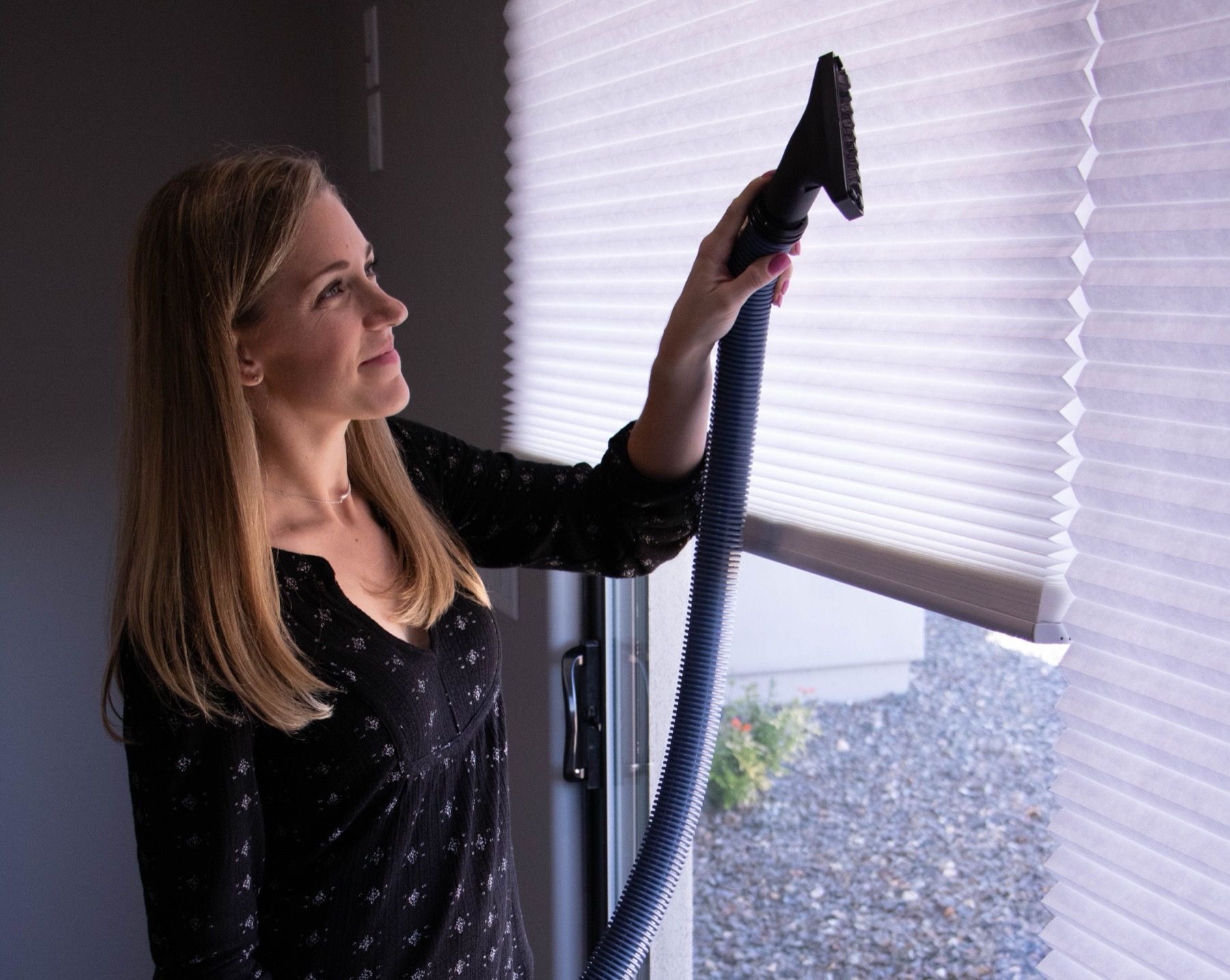 A woman at a window running a central vacuum hose up to the top of a pleated blind.