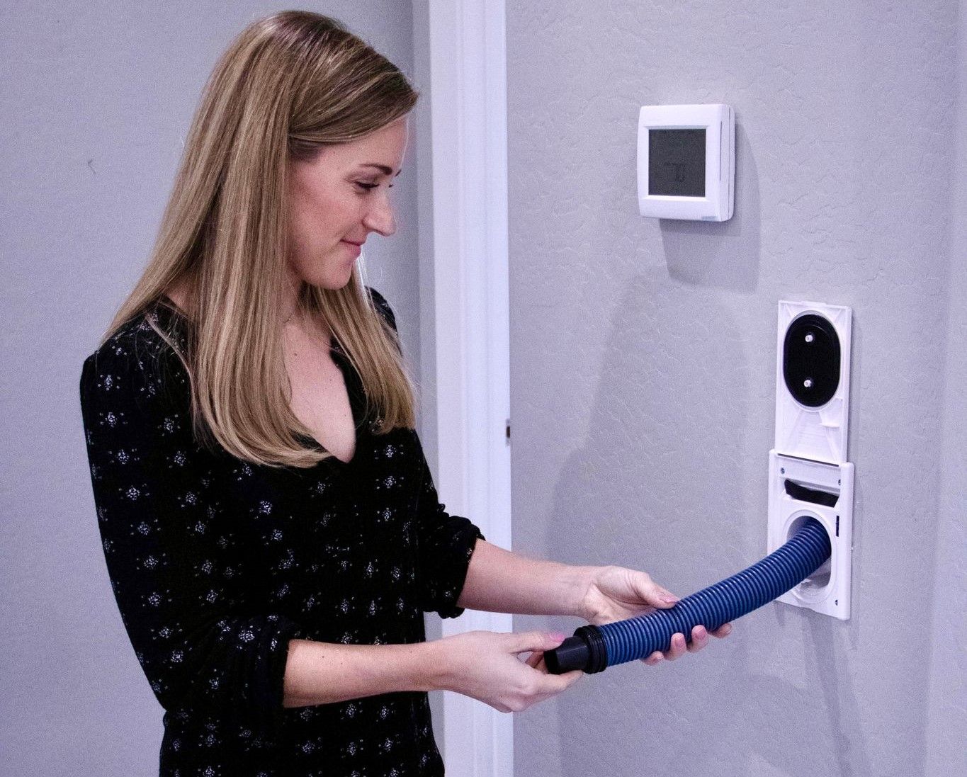 A woman standing at a wall in a hallway, holding the cuffed end of a blue central vacuum hose at an open wall inlet.