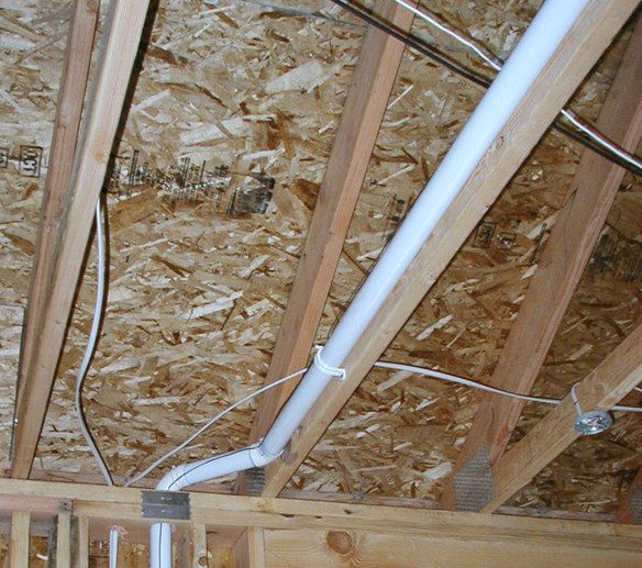 Central vacuum tubing roughed into an unfinished house: white pipe running up between open ceiling joists, a branch elbow dropping off it, and low-voltage wire stapled along the framing.