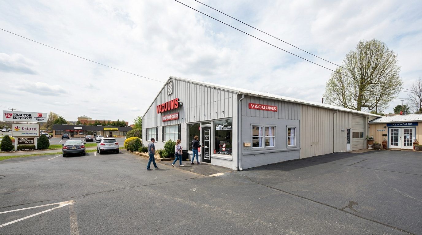 The storefront: a long single-storey building with red VACUUMS signs on the gable end and along the side wall, glass entrance doors at the front, a parking lot with cars in it, and a retail strip further back.